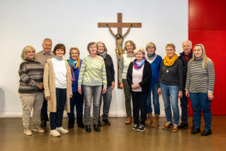 Männer und Frauen stehen zu einem Gruppenbild zusammen, in der Mitte des Bildes hängt ein Kreuz an der Wand